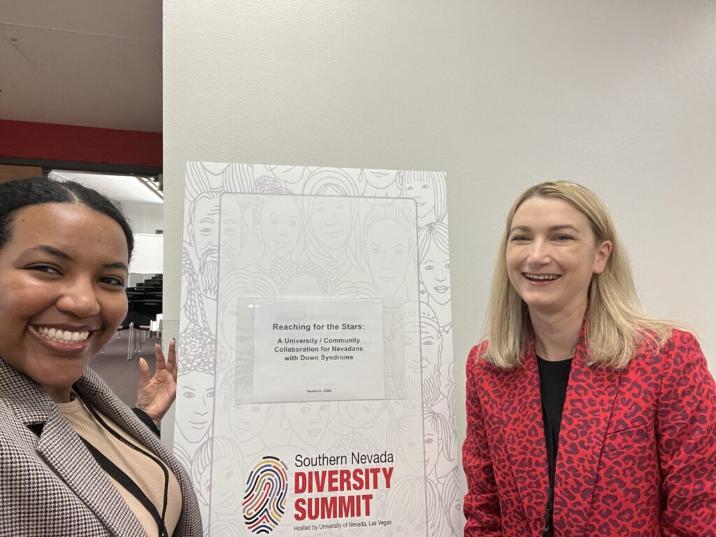Samrawit Misiker (left) and Kate Martin (right) are standing on each side of a Southern Nevada Diversity Summit sign that states, "Reaching the Stars: A University / Community Collaboration for Nevadans with Down Syndrome." Samrawit is wearing a brown and white patterned blazer and Kate is wearing a red and black patterned blazer.